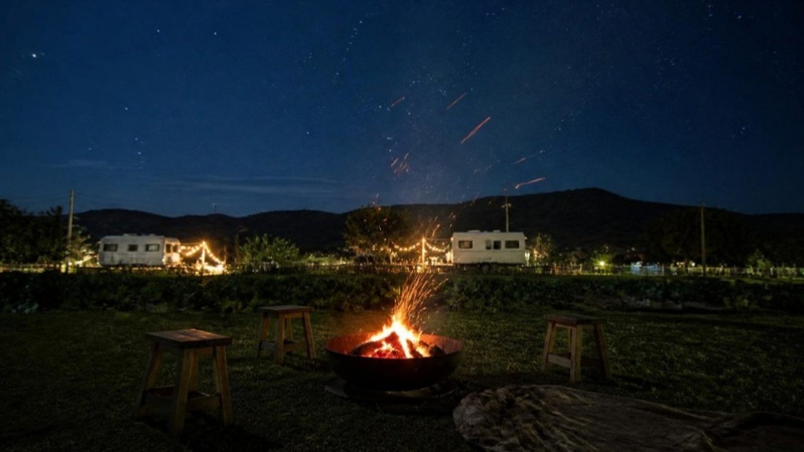 示意圖：夜裡的農場草地上，一盆營火旁放著幾張木凳與毛毯，火星升向星空，遠處是白色露營車與燈串。