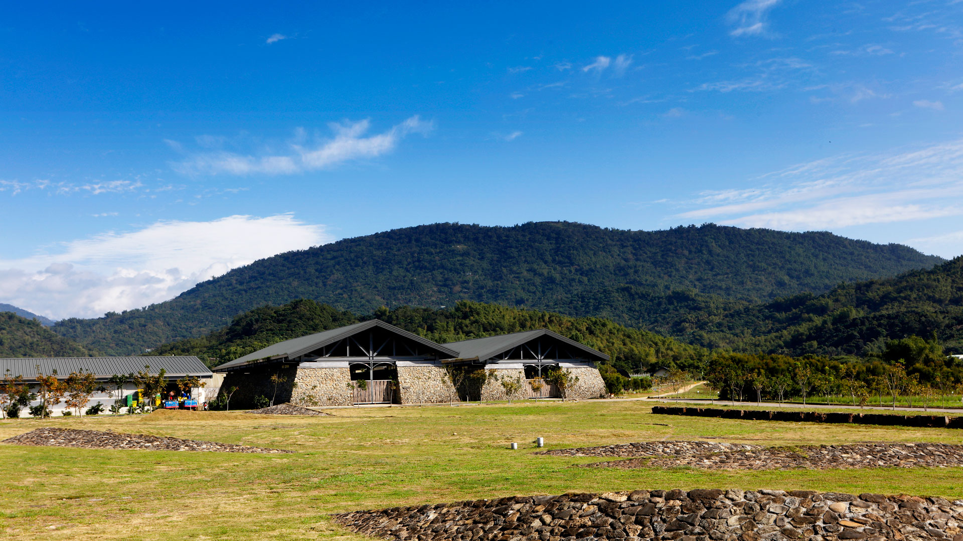 石牆斜屋頂的建築坐落在草地上，後面是一整片青山和藍天。